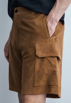 Brązowe szorty typu cargo zamszowe z kieszenią na klapę, noszone przez osobę z jedną ręką w przedniej kieszeni na jednolitym tle.