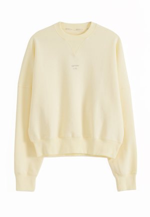 Vaaleankeltainen pyöreäkauluksinen collegepaita, jossa on pitkät hihat ja pieni, keskitetty kirjailtu logo rinnassa.