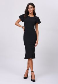 Robe noire ajustée avec des manches courtes à volants, longueur au genou et bas évasé. Portée avec des chaussures à talons hauts noires et des boucles d'oreilles en argent en forme de créole.
