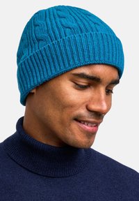 Turquoise knit beanie with a textured cable pattern, featuring a ribbed bottom edge. Worn with a dark turtleneck sweater.