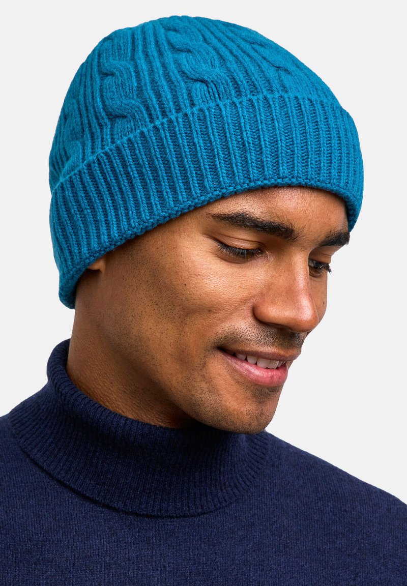 Turquoise knit beanie with a textured cable pattern, featuring a ribbed bottom edge. Worn with a dark turtleneck sweater.