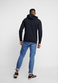 Muž v černé mikině s kapucí, modrých vykasaných džínách a černobílých teniskách, stojící na jednobarevném světle šedém pozadí.