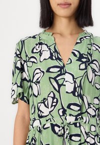Grønn blomstret kjole med korte ermer, med V-hals og strammesnor i midjen, designet med svart-hvite blomsterdesign.