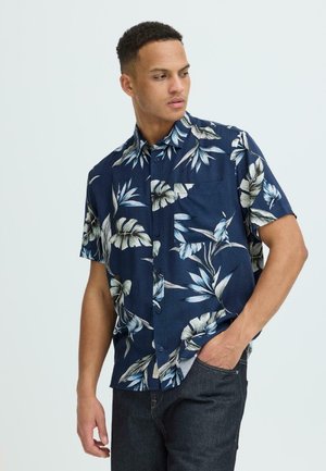 Homme portant une chemise à manches courtes bleu marine avec un imprimé de feuilles tropicales blanches et bleu clair, la main dans la poche d'un jean foncé, regardant vers la gauche.