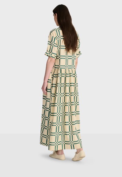 Femme aux longs cheveux bruns portant une robe maxi crème avec un motif à carreaux verts et des chaussures beige, debout devant un fond uni.