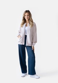 Jasnoróżowa kurtka na zamek z prążkowanymi mankietami, biała koszulka i granatowe spodnie szerokiego kroju. Modelka w białych trampkach, ręce w kieszeniach kurtki.