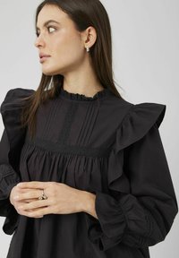 Blouse noire avec épaules à volants, col montant et bordure en dentelle décorative. Présente un tissu froncé et une patte avec des plis verticaux.