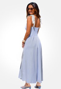 Blau-weiß gestreiftes Maxi-Kleid mit gerafftem Oberteil, gebundenen Trägern und fließendem Stoff. Getragen mit blauen High-Heels und Sonnenbrille.