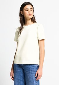 Haut crème à manches courtes avec un col rond, présentant des détails légèrement bouffants sur les manches et un tissu lisse et texturé.
