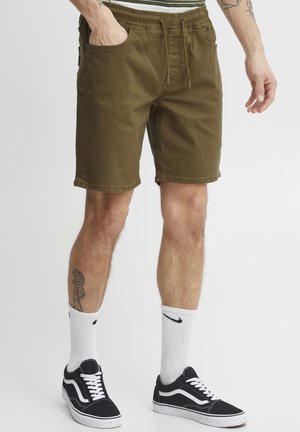 Shorts vert olive avec poches latérales et taille élastique. Portés avec des baskets noires et des chaussettes blanches avec un logo.