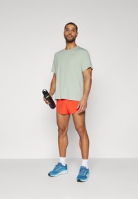Sportshorts i lys oransje, en lys grønn t-skjorte, og blå atletisk sko, holder en svart vannflaske. Rette linjer og moderne passform.