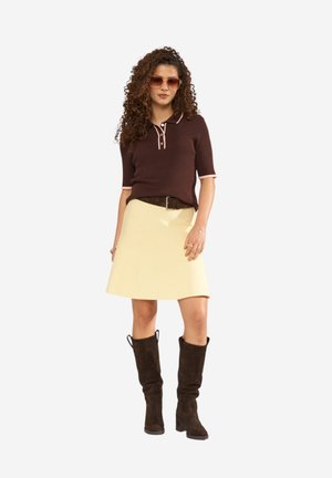 Femme aux cheveux bouclés portant un polo marron, une jupe beige, une ceinture marron, de grandes bottes marron et des lunettes de soleil sur fond blanc.