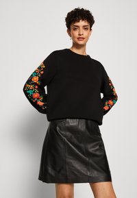 Sudadera negra con bordados florales coloridos en las mangas, combinada con una falda de cuero negra. Tejido suave y diseño de corte relajado.