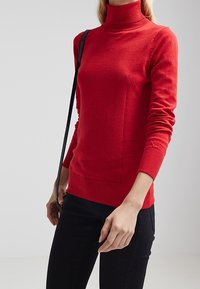 Roter Rollkragenpullover mit langen Ärmeln, geripptem Saum und Bündchen, aus weichem Strickstoff, kombiniert mit schwarzen Hosen und einer Umhängetasche.