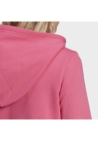 Mikina s kapucí v jasně růžové barvě, vyznačující se měkkou tkaninou a žebrovanými manžetami. Pohled zezadu zdůrazňuje tvar a konstrukci kapuce.