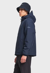 Homme portant une veste à capuche bleu marine et un chapeau bob assorti, debout de profil sur un fond clair uni.
