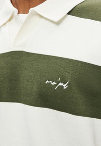 Valge ja oliivroheliste triipudega polosärk, millel on kraega kaelus ja valge tikitud logo. Pehme, tekstuuriga kangas.