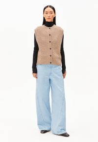 Beige gebreid vest met een hoge kraag, knoopsluiting aan de voorkant en geribbelde zoom, gedragen over een zwarte top met lange mouwen en gecombineerd met lichtblauwe broek met wijde pijpen.