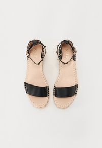 Sandales en cuir noir avec une large sangle, des clous décoratifs au niveau de la cheville, une semelle intérieure beige et une semelle en jute tressé. Fermeture à boucle réglable.
