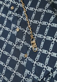 Chemise bleu marine avec un motif géométrique blanc, dotée de boutons dorés et d'un détail en chaîne avec un fermoir, mettant en valeur un tissu texturé.