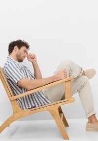 Wooden lounge chair with a slatted design, featuring a light brown finish. The subject wears a blue and white striped short-sleeve shirt and light trousers.