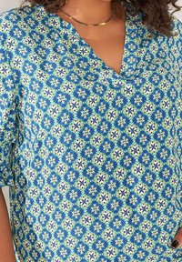 Blusa in denim con scollo a V, maniche corte e un motivo geometrico floreale blu e verde su uno sfondo chiaro.