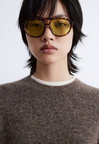 Brauner, flauschiger Pullover mit rundem Ausschnitt, getragen unter einer Schildkrötenbrillen mit gelben Gläsern und kantigem Design.