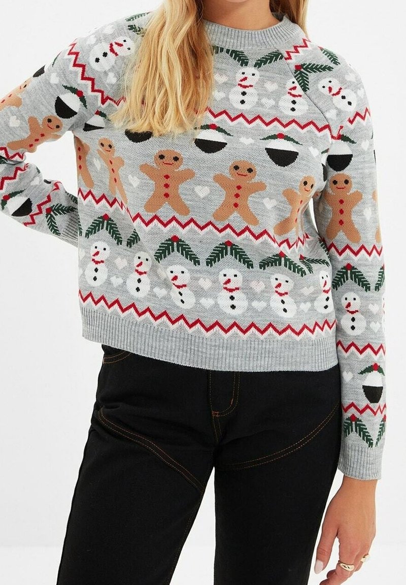 Femme portant un pull de Noël gris orné de bonshommes en pain d'épice, de bonhommes de neige, de feuilles de houx et de motifs en zigzag rouges.