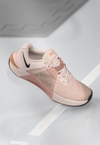Lichtroze sportschoen met een gestructureerde mesh bovenzijde, zwarte Nike swoosh en roze accenten. Versterkte hak en rubberen buitenzool voor grip.