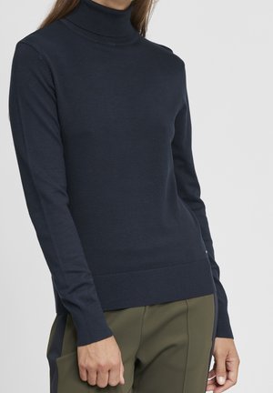 Femme portant un pull à col roulé moulant bleu marine foncé et un pantalon vert olive sur un fond clair uni.
