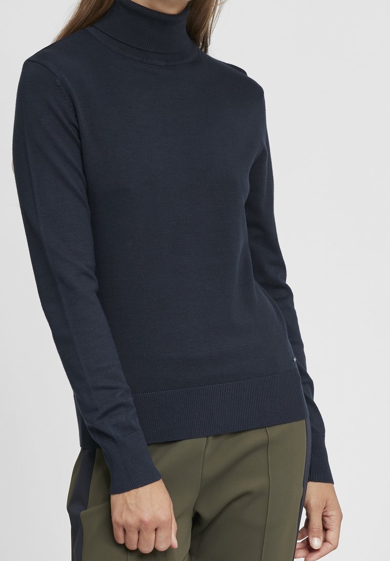 Femme portant un pull à col roulé moulant bleu marine foncé et un pantalon vert olive sur un fond clair uni.