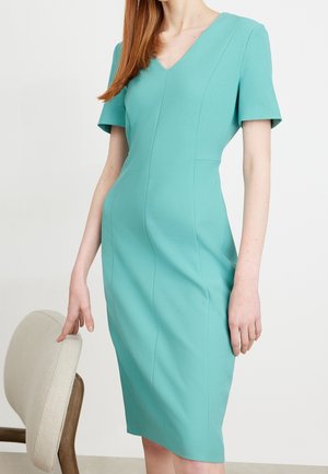 Nainen pukeutunut polvipituiseen turkoosiin mekkoon, jossa on lyhyet hihat ja V-aukko, seisoo beigen pehmustetun tuolin vieressä.