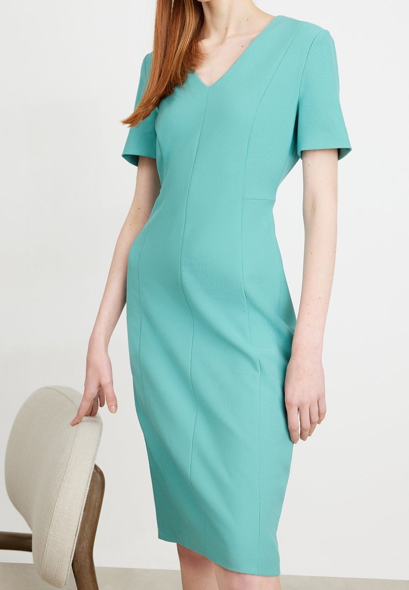 Femme portant une robe turquoise mi-longue à manches courtes et col en V, debout à côté d'un fauteuil beige rembourré.