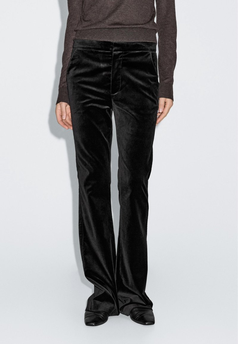 Personne portant un pantalon droit en velours noir, un haut à manches longues marron foncé et des chaussures noires à bout pointu, debout devant un fond clair uni.