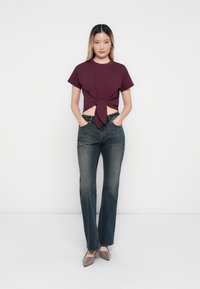 Bordeaux-kleurige cropped t-shirt met een knoopdetail aan de voorkant, gecombineerd met donkerblauwe flare jeans. Het model draagt lichtbruine puntige hakken.