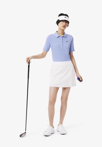 Polo azul claro de manga corta con tres botones, acompañado de una falda blanca. Los accesorios incluyen una visera blanca y un palo de golf.