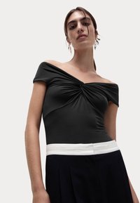 Sort off-the-shoulder bodysuit med tvistet stofdetalje ved brystet, parret med en hvid talje og mørke plisserede bukser.