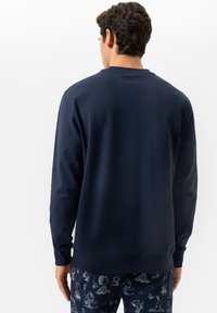 Un homme avec des cheveux courts et bouclés porte une chemise à manches longues bleu marine et un pantalon bleu marine avec des imprimés de personnages de dessin animé blancs, se tenant devant un fond blanc.