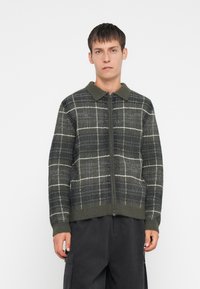 Cardigan com fecho éclair em padrão xadrez verde e cinza, com colarinho, tecido texturizado e parte inferior canelada. Os detalhes incluem padrões brancos em grelha e um zíper suave.