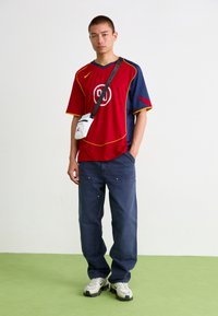 Rød og blå mesh trøje med et cirkulært logo, parret med løse mørkeblå bukser og hvide sneakers. Lille hvid crossbody taske.