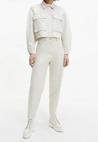 Zkrácená béžová bunda se dvěma kapsami na hrudi a štítkem; v kombinaci se světle béžovými tapered kalhotami a bílými šněrovacími platformovými botami.