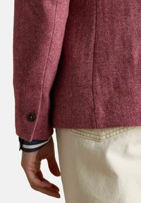 Ruka spočívajúca na boku, oblečená v krémových nohaviciach a textúrovanom ružovom sakku s gombíkovým manžetom cez pruhovanú rukáv.