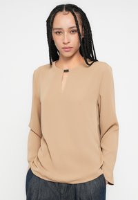 Bluză bej cu mâneci lungi, cu decolteu rotund și detaliu cheie în față. Confecționată dintr-un material neted; prezintă un drapaj subtil pe părțile laterale.