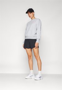 Felpa grigia con polsini a coste, abbinata a pantaloni corti neri e sneakers bianche, completata da un cappellino nero con scritte in rilievo.