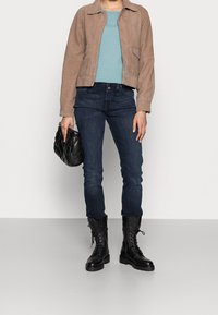Veste en daim beige, haut texturé bleu clair sarcelle, jean slim bleu foncé, et bottes de combat noires à lacets. Sac à main matelassé noir à la main.