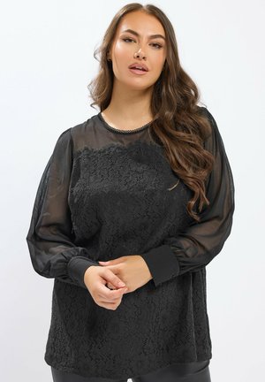 Kvinna med långt vågigt hår iförd en svart spetsblus med genomskinliga ballongärmar, stående med händerna knäppta mot en enkel vit bakgrund.