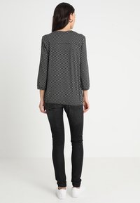 Frau mit langen dunklen Haaren, die eine schwarz-weiße gemusterte Bluse, schwarze enge Jeans und weiße Sneakers trägt, steht mit dem Rücken zur Kamera.
