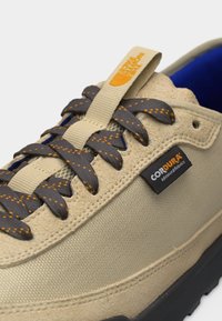 Μπεζ αθλητικό παπούτσι με μαύρα και πορτοκαλί κορδόνια με πουά, ετικέτα από ύφασμα CORDURA και κίτρινη ταινία με λογότυπο στο γλώσσο.