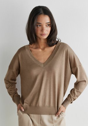 Frau mit schulterlangem dunklem Haar, die einen lockeren braunen Pullover mit V-Ausschnitt und beigefarbene Hosen trägt, steht vor einer schlichten weißen Wand.