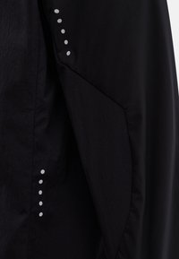 Veste noire avec un tissu texturé orné de pois blancs contrastants le long de la couture, présentant un design minimaliste et des formes angulaires.